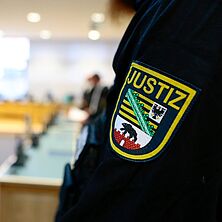 Ärmel mit Logo eines Justizbeamten im Vordergrund, dahinter verschwommen der Gerichtssaal